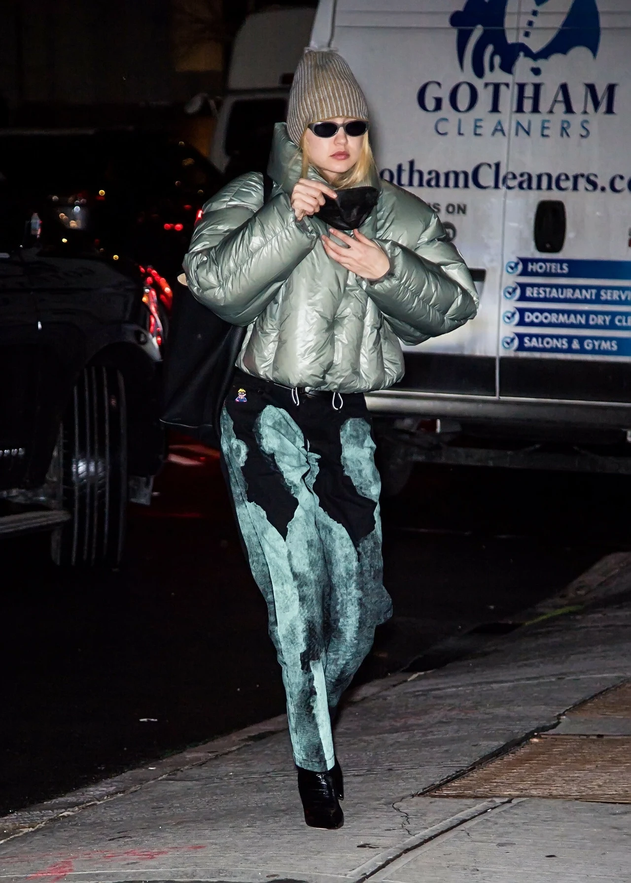Gigi Hadid w krótkiej kurtce puchowej Kobieta ubrana w srebrną kurtkę puchową, zieloną czapkę i okulary przeciwsłoneczne idzie chodnikiem nocą, trzymając telefon i kawę, w tle pojazd dostawczy z napisem Gotham Cleaners.