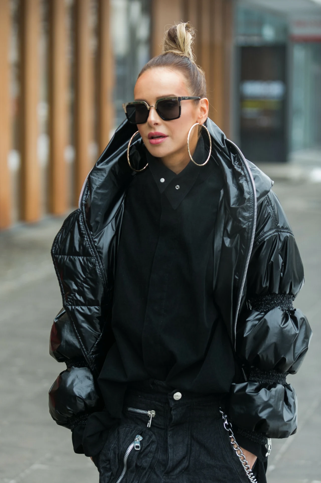 Magdalena Mielcarz w oversize'owej, czarnej kurtce puchowej Kobieta w czarnych, nowoczesnych ubraniach, ubrana w połyskującą puchową kurtkę, duże kolczyki koła i ciemne okulary przeciwsłoneczne, stoi na tle miejskiego chodnika.