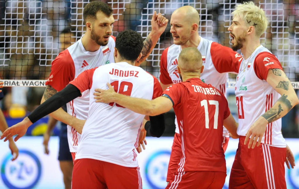 Norbert Huber z kolegami z siatkarskiej reprezentacji Polski Grupa siatkarzy ubranych w biało-czerwone stroje świętuje wspólnie zdobycie punktu na boisku, widoczna siatka oraz fragment publiczności w tle.