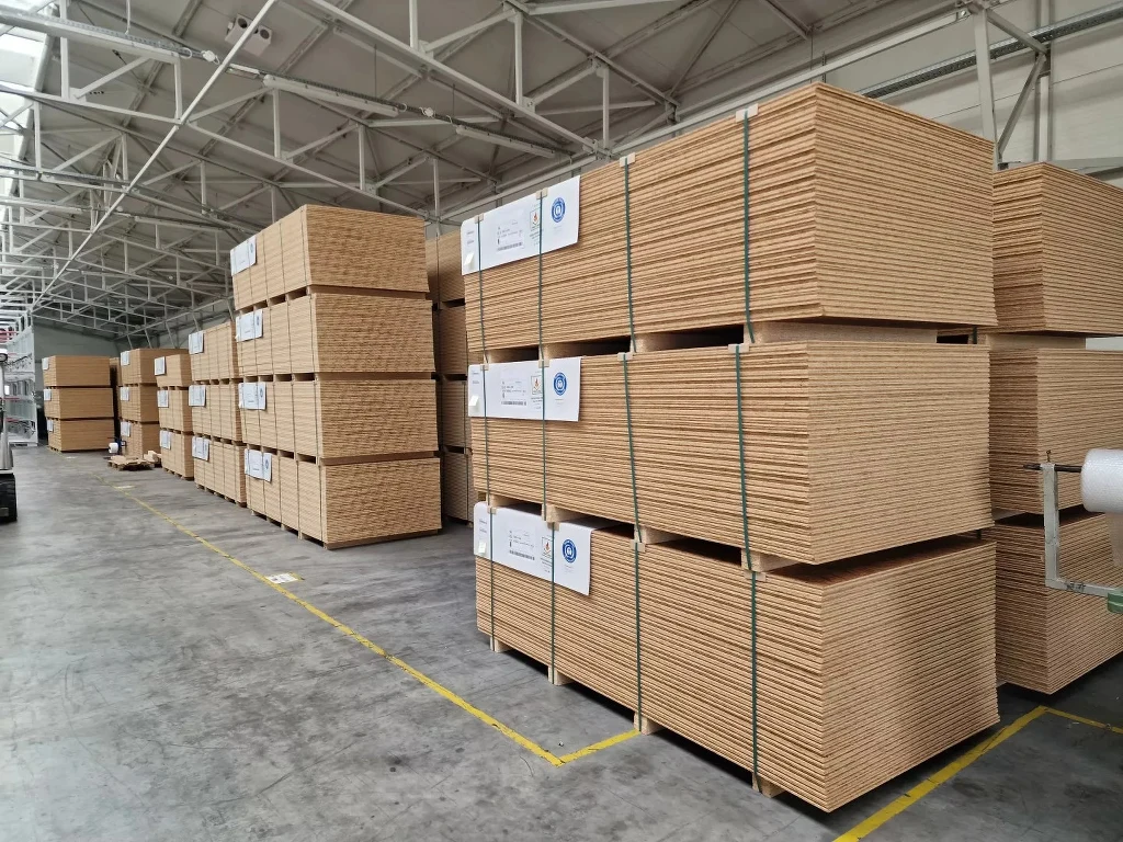 Stosy płyt drewnopochodnych ustawione równo na paletach w dużym, dobrze oświetlonym magazynie przemysłowym z betonową posadzką i metalową konstrukcją dachu.