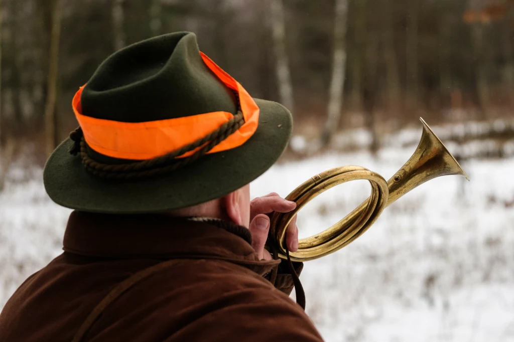 Czy myśliwi powinni mieć wstęp do szkół? Mężczyzna w zielonym kapeluszu z pomarańczową wstążką gra na rogu myśliwskim, stoi w zimowym, leśnym otoczeniu.