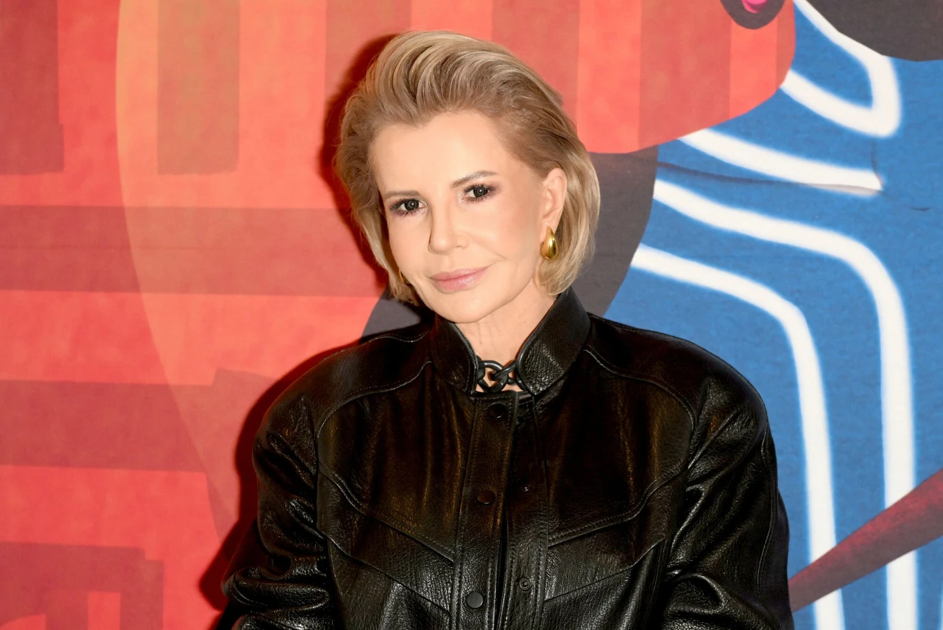 Joanna Racewicz Kobieta z krótkimi blond włosami ubrana w czarną skórzaną kurtkę stoi na tle kolorowej, abstrakcyjnej grafiki z czerwonymi i niebieskimi elementami.