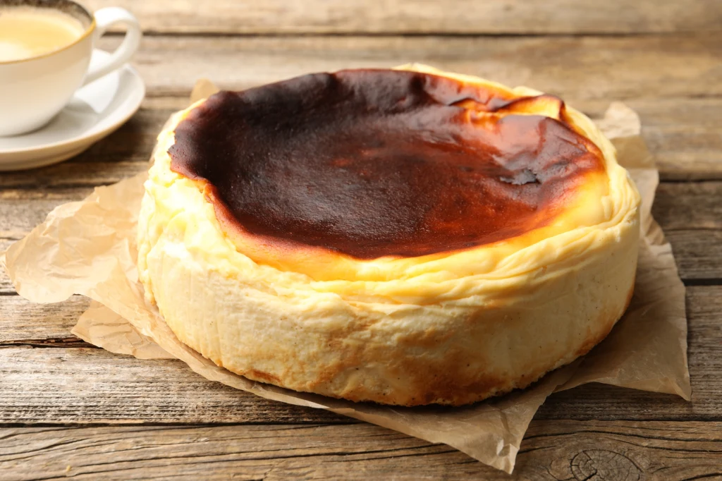 Na co zwrócić uwagę podczas korzystania z papieru do pieczenia? Sernik baskijski z charakterystycznie przypieczonym, ciemnobrązowym wierzchem, ustawiony na pergaminie na drewnianym stole, obok filiżanki kawy.