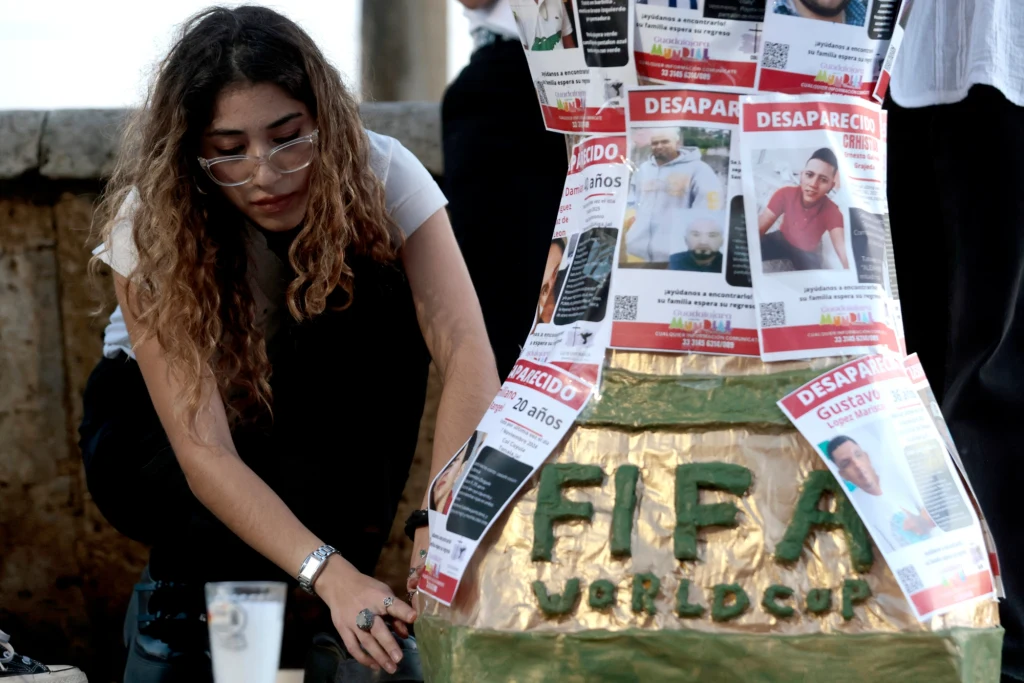 Demonstracja studentów w Guadalajarze Kobieta z długimi włosami stoi obok makiety trofeum FIFA World Cup, która jest oklejona licznymi plakatami osób zaginionych. Wokół panuje poważna atmosfera, a plakaty zawierają zdjęcia oraz informacje o zaginionych osobach.