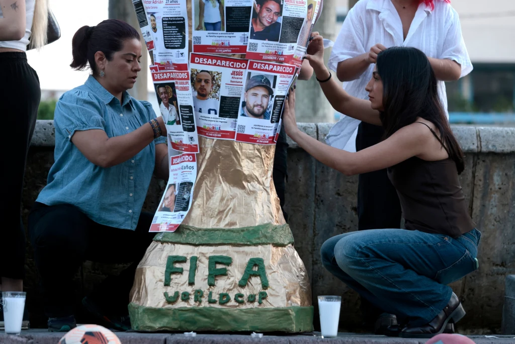 Demonstracja studentów w Guadalajarze Dwie kobiety przyklejają plakaty ze zdjęciami zaginionych osób na dużym modelu pucharu świata FIFA, obok ustawiono zapalone świece, widoczna jest również piłka nożna w dolnej części obrazu.