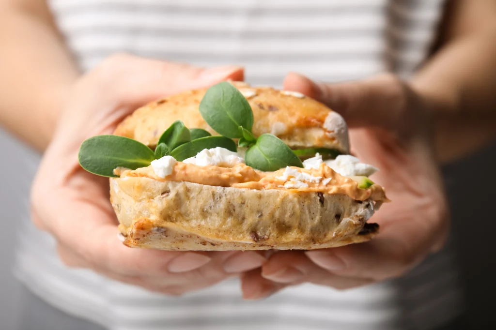 Kanapka z chrupiącą bułką, kremowym serkiem, świeżymi liśćmi szpinaku i apetyczną pastą, trzymana w dwóch dłoniach na tle pasa w paski.