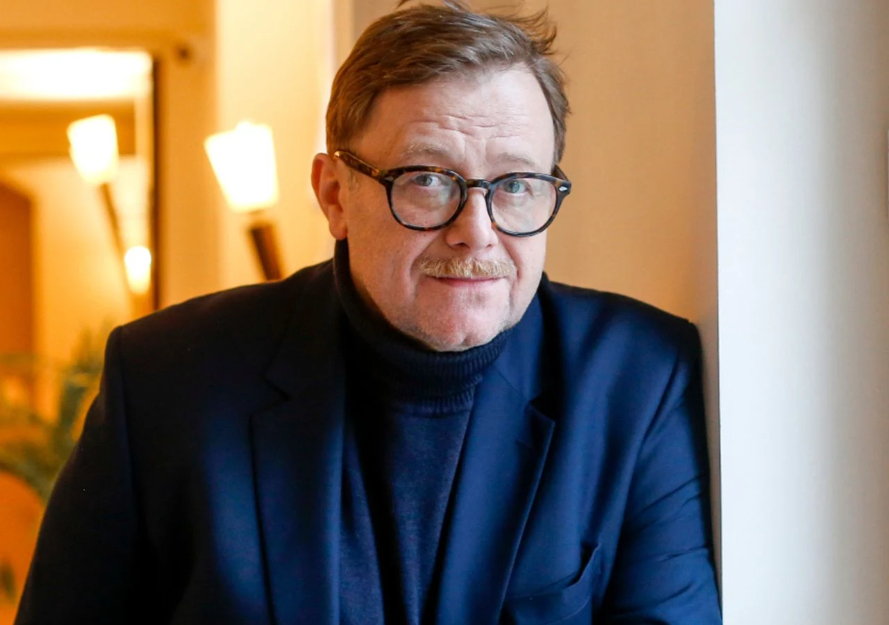 Olaf Lubaszenko Mężczyzna w okularach i ciemnym garniturze opiera się o jasną ścianę, w tle ciepłe światło lamp i rozmyte wnętrze.