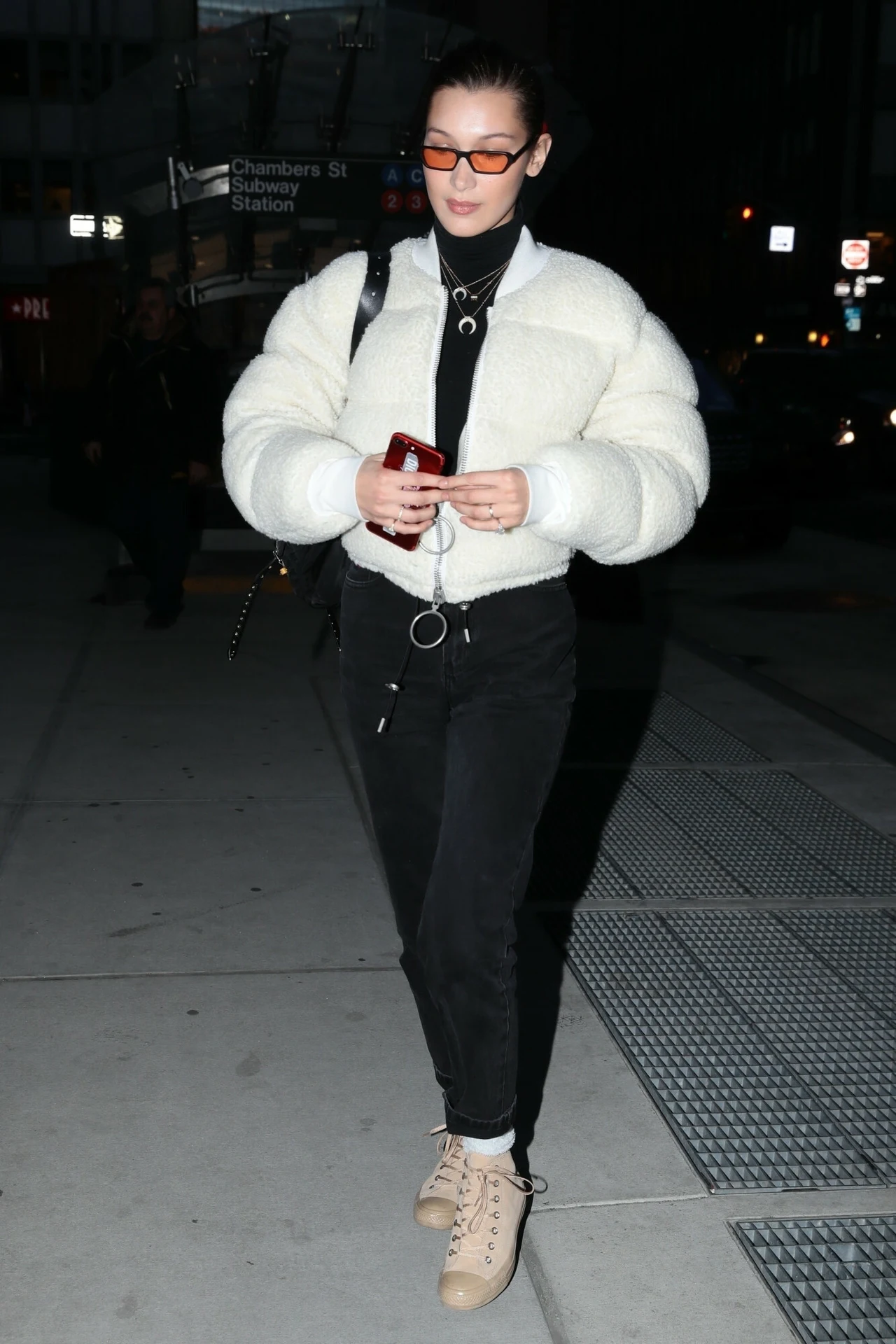 Bella Hadid w białej, krótkiej kurtce Młoda kobieta w białej puchowej kurtce, czarnych spodniach i jasnych butach idzie ulicą miasta nocą, trzymając telefon. Ma na sobie ciemne okulary i czarne ubrania pod kurtką. W tle widoczne są rozświetlone miejskie światła i znaki drogowe.