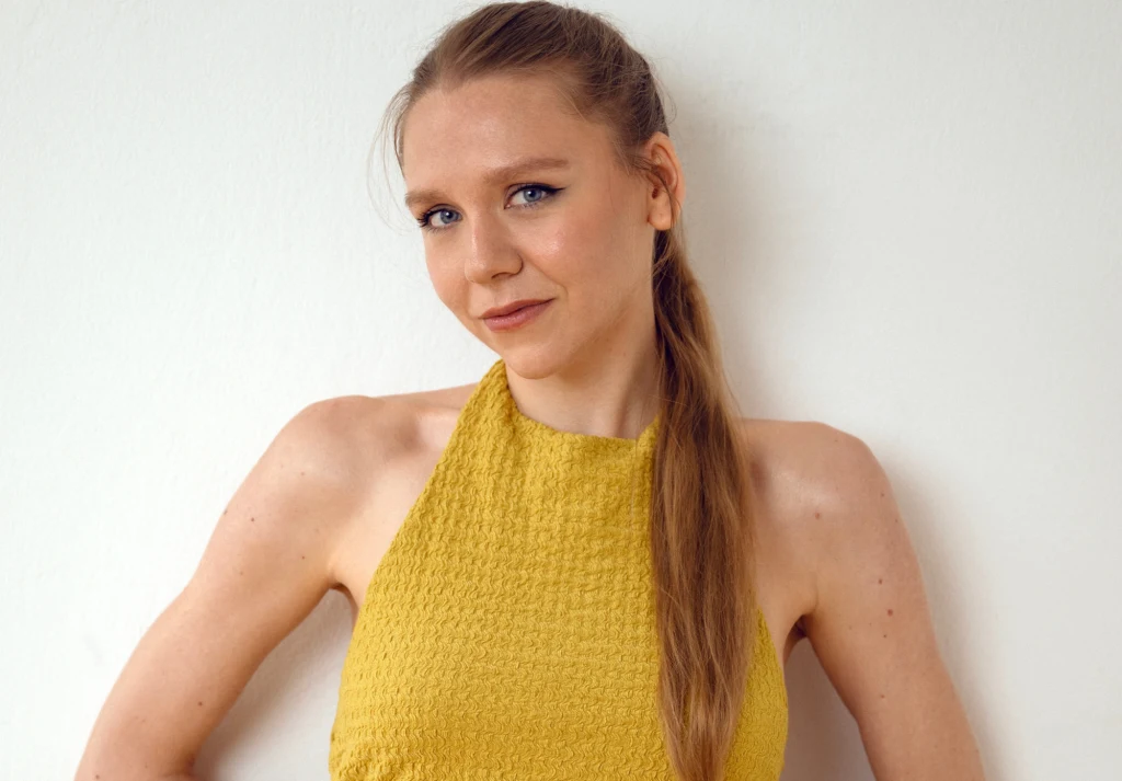 Maja Staśko, autorka książki "Siostry w bólu. Historie o endometriozie" Kobieta o jasnych włosach spiętych w koński ogon, ubrana w żółty top z fakturą, stoi na tle białej ściany i uśmiecha się delikatnie do obiektywu.