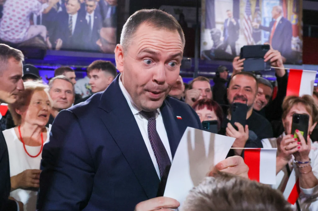 Prezydent RP - Karol Nawrocki Mężczyzna w garniturze z polską flagą na krawacie trzyma dokument i patrzy na niego z zaskoczoną miną. Wokół zgromadzeni ludzie, część z nich robi zdjęcia telefonami komórkowymi, w tle widoczne flagi Polski i duże plakaty przedstawiające spotkania poli...