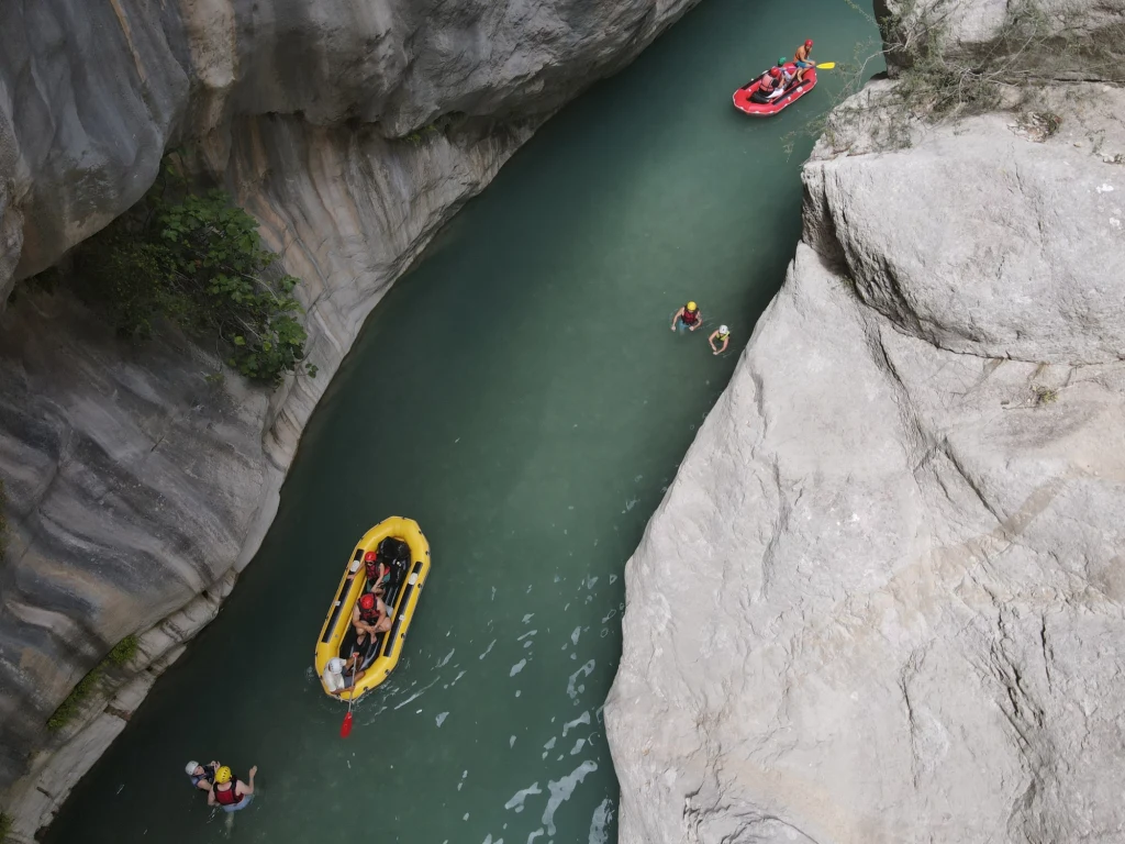 Dwie grupy osób płyną na pontonach przez wąski, skalisty kanion, towarzyszą im osoby płynące w wodzie. Strome ściany kanionu otaczają spokojnie płynącą rzekę.