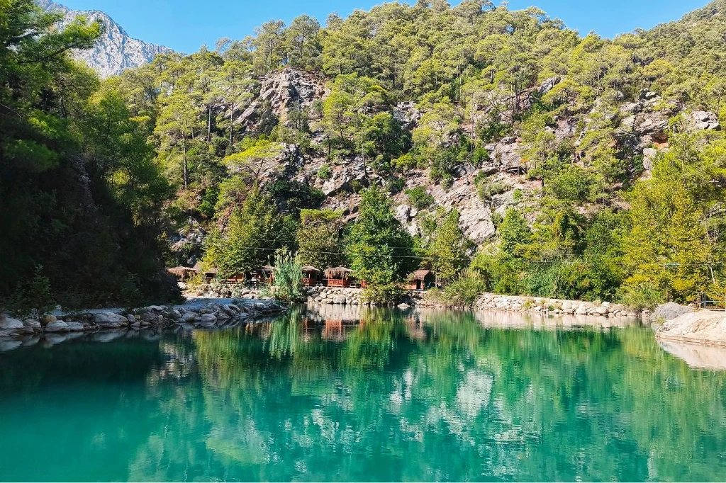 Turkusowe jezioro otoczone gęstym, zielonym lasem i stromymi skałami, odbijające w wodzie drzewa oraz fragmenty zabudowań schowanych pośród przyrody, w oddali widoczne góry.