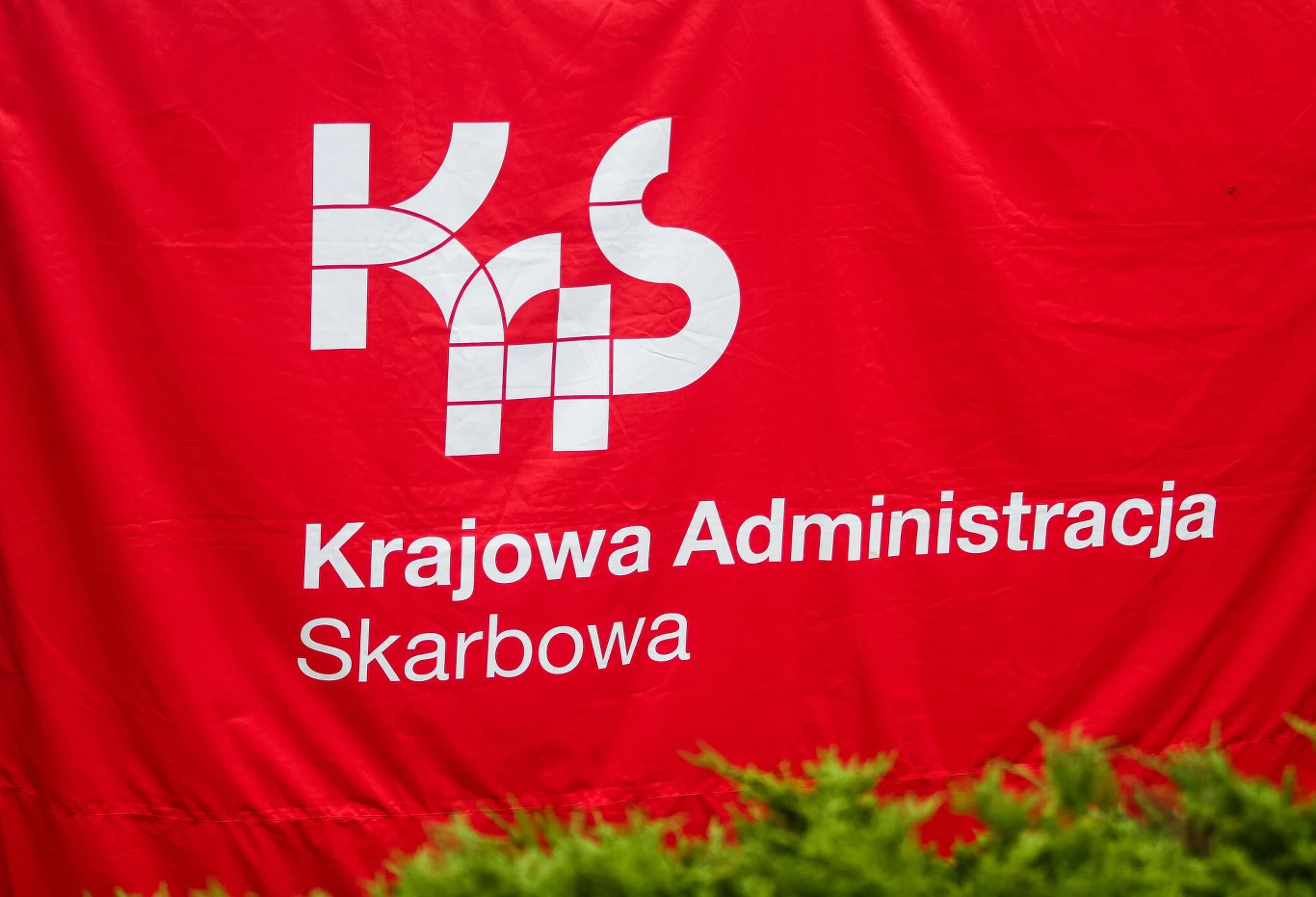 KAS wykryła nieprawidłowości w CIT spółki farmaceutycznej (zdj. ilustracyjne) Logo Krajowej Administracji Skarbowej na czerwonym tle, pod nim widoczny biały napis z nazwą instytucji, w dolnej części zielone liście krzewu.