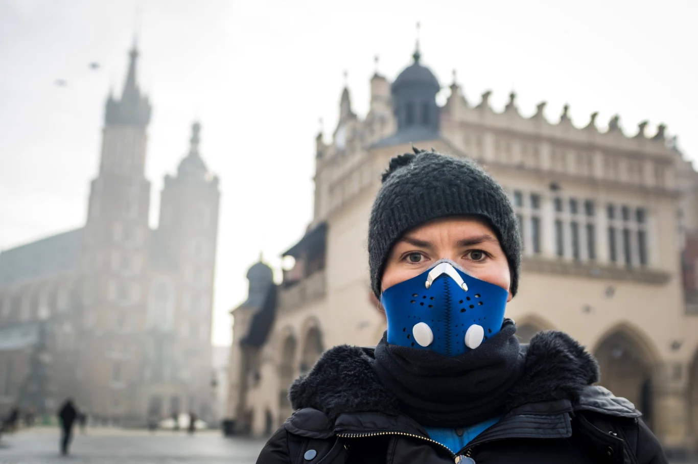 Większość Polaków odczuwa problemy ze zdrowiem w wyniku smogu - wskazuje najnowsza ankieta Osoba nosząca maskę antysmogową stojąca na tle charakterystycznych, zabytkowych budynków rynku w Krakowie, w zimowej czapce i kurtce, widoczne zamglenie lub smog wokół.