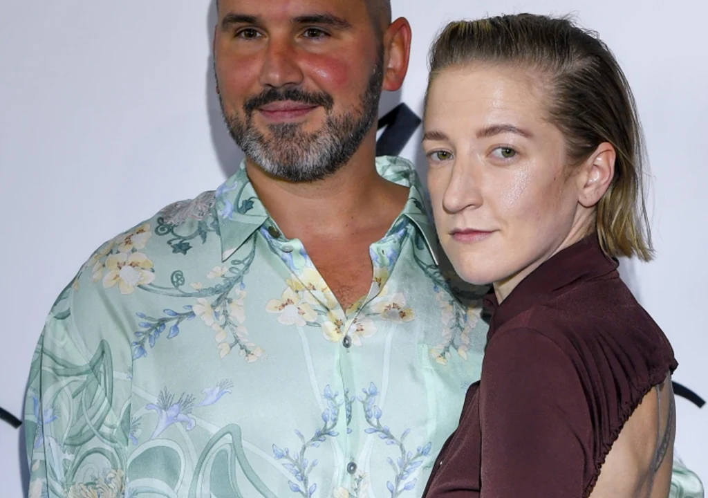 Piotr Szeląg i Lara Gessler Dwie osoby pozują razem na tle jasnej ścianki z czarnym napisem, mężczyzna w jasnej, wzorzystej koszuli oraz kobieta w bordowym stroju z tatuażem na ramieniu.