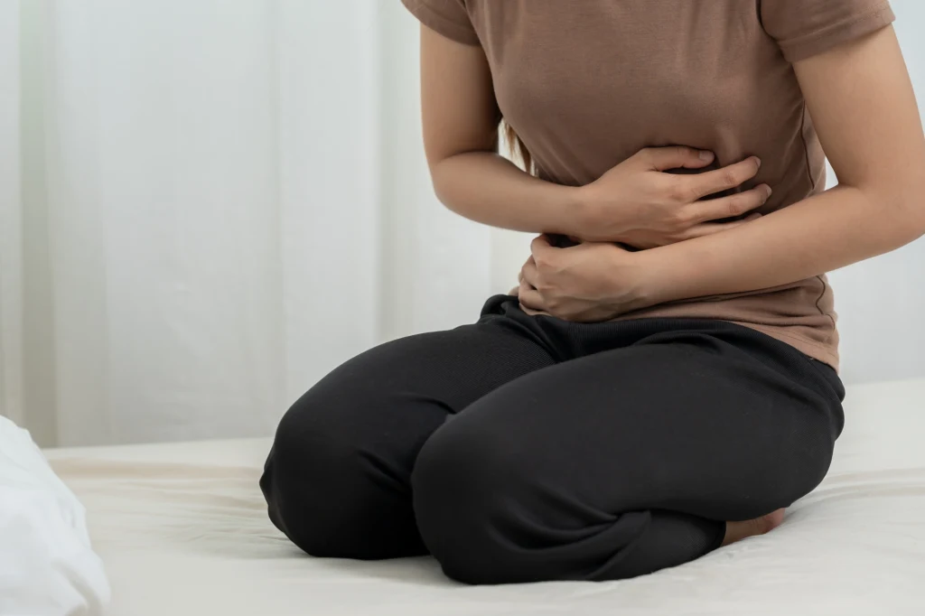 Podczas ataku ból może być tak silny, że utrudnia chodzenie Osoba w brązowej koszulce i czarnych spodniach siedzi na łóżku z rękami przyłożonymi do brzucha, sugerując dolegliwości bólowe lub dyskomfort w okolicy brzucha.