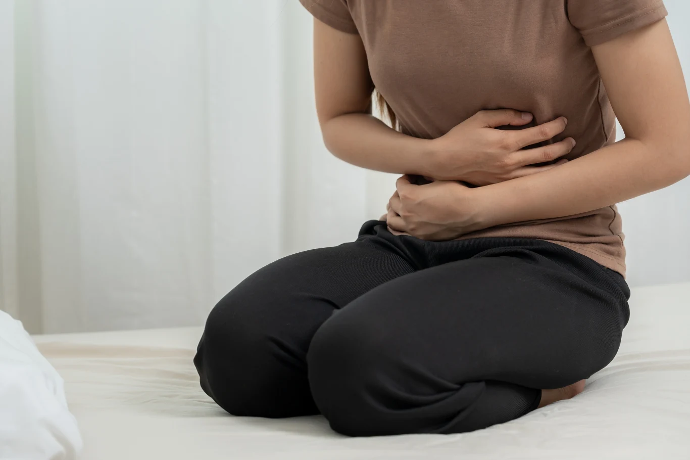 Podczas ataku ból może być tak silny, że utrudnia chodzenie Osoba w brązowej koszulce i czarnych spodniach siedzi na łóżku z rękami przyłożonymi do brzucha, sugerując dolegliwości bólowe lub dyskomfort w okolicy brzucha.