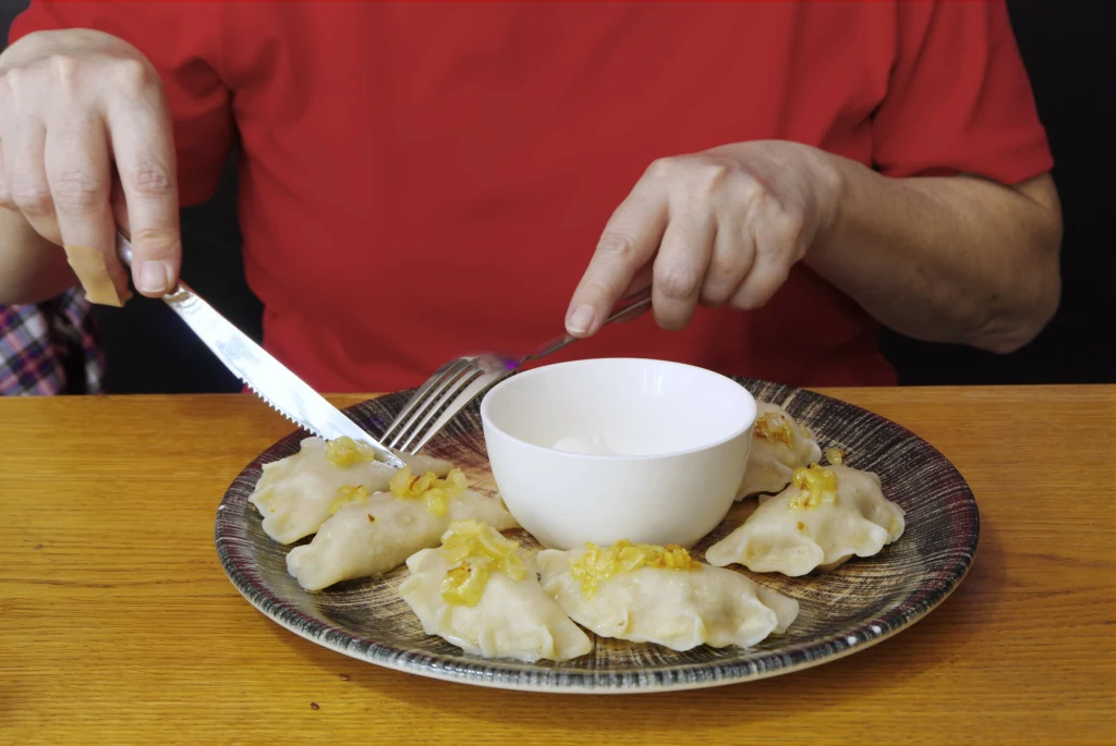 Gdzie na pyszne pierogi w Gdyni? Talerz z pierogami udekorowanymi skwarkami oraz miseczka ze śmietaną pośrodku, na pierwszym planie ręce osoby w czerwonej koszulce trzymające widelec i nóż.