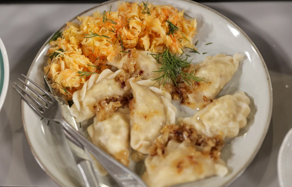 Gdzie w Trójmieście zjeść pierogi? Porcja pierogów polanych zeszkloną cebulką podana z surówką z marchwi i kapusty, dekorowana świeżym koperkiem, na białym talerzu z widelcem i nożem.