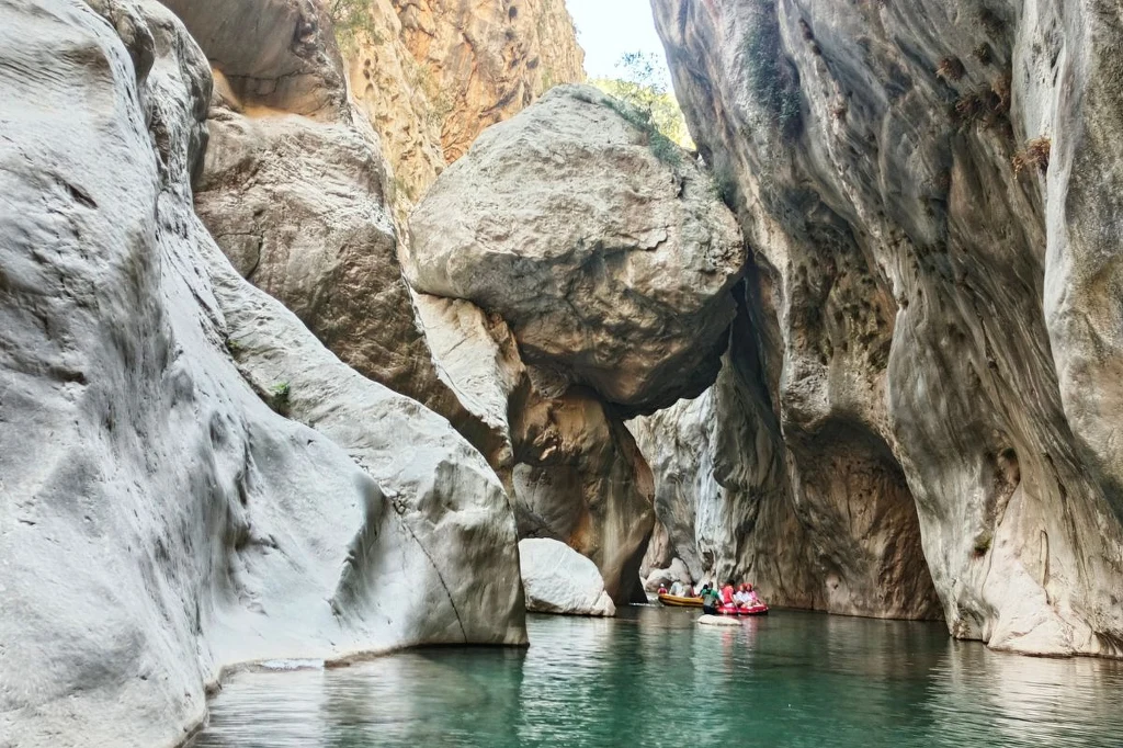 Wąwóz z wysokimi, stromymi ścianami skalnymi, przez który przepływa rzeka. Grupa osób na pontonach płynie wśród malowniczych skał, jasny kolor kamieni kontrastuje z zieloną wodą.