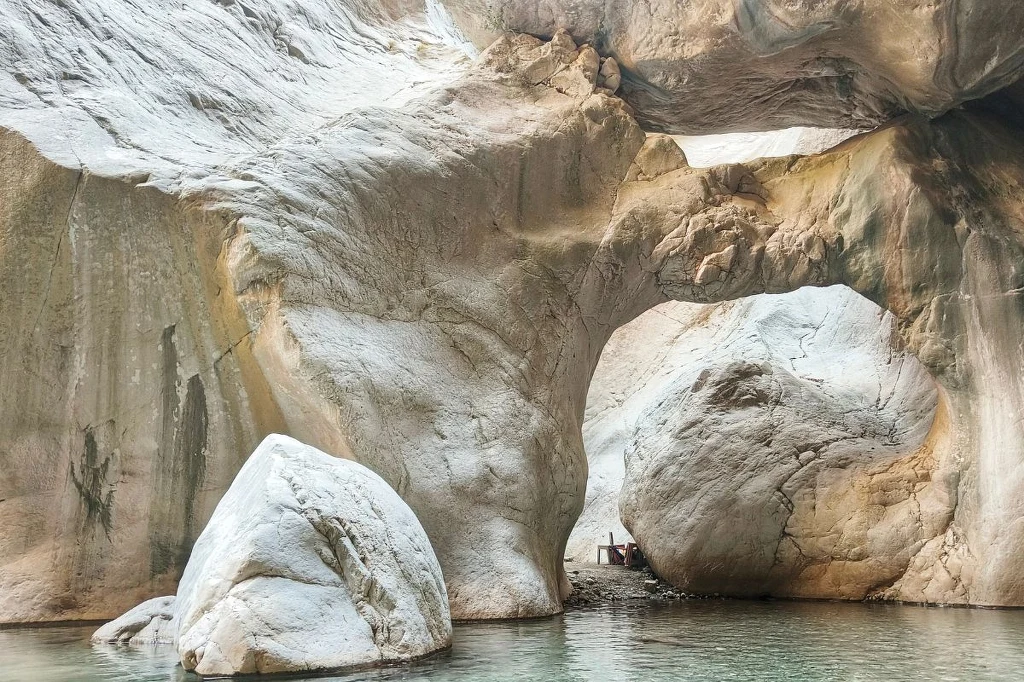 Ogromne, jasne skały z naturalnym łukiem skalnym nad spokojną wodą, w tle widać prześwit światła i niewielkie drewniane pomosty przy dużych głazach.