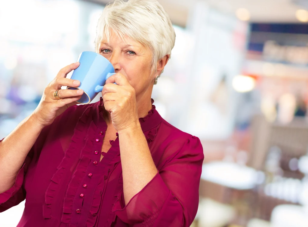 O jakiej porze najlepiej pić kawę? Starsza kobieta o krótkich, siwych włosach ubrana w ciemnoróżową bluzkę, trzymająca niebieski kubek i pijąca napój, z rozmytym tłem jasnej kawiarni.
