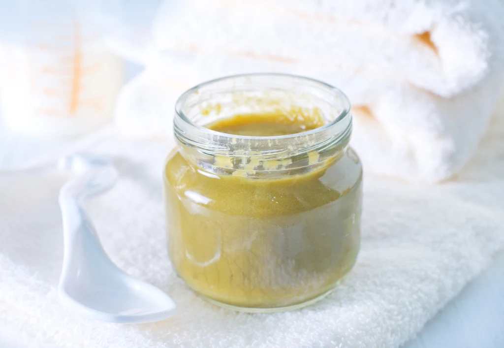 Szklany słoik z zieloną papką lub puree ustawiony na białym ręczniku obok plastikowej łyżeczki, w tle rozwinięty ręcznik kąpielowy i częściowo widoczny pojemnik miarowy.