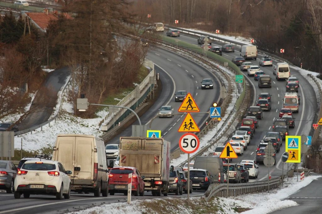 Zatłoczona droga szybkiego ruchu w zimowej scenerii, na obu jezdniach widoczne są liczne samochody jadące w obu kierunkach, znaki drogowe informują o ograniczeniu prędkości i przejściu dla pieszych, pobocza pokryte cienką warstwą śniegu.
