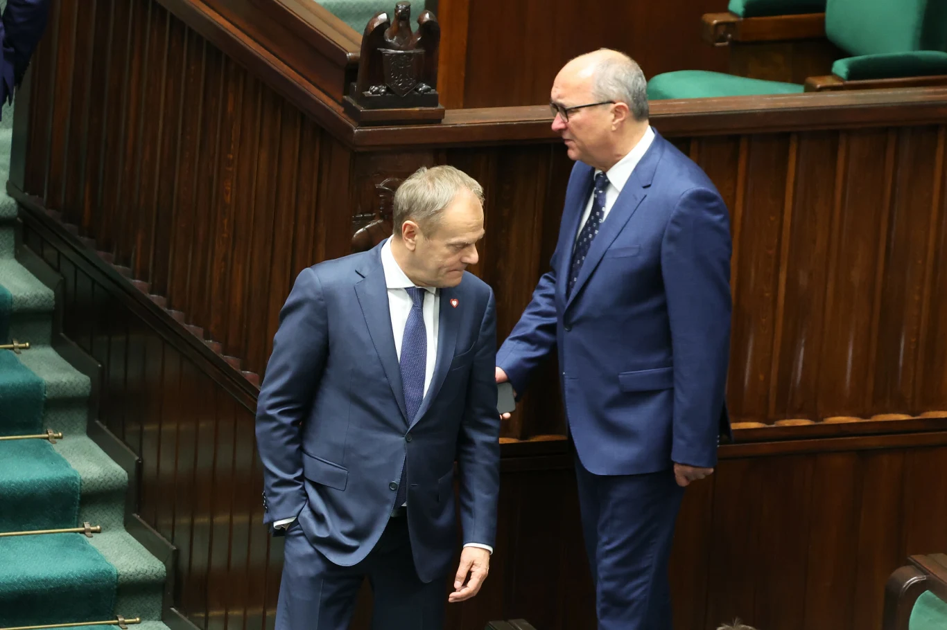 Dwóch mężczyzn w garniturach rozmawia na schodach w sali sejmowej, wokół drewniane barierki i zielone obicia foteli, atmosfera formalna.