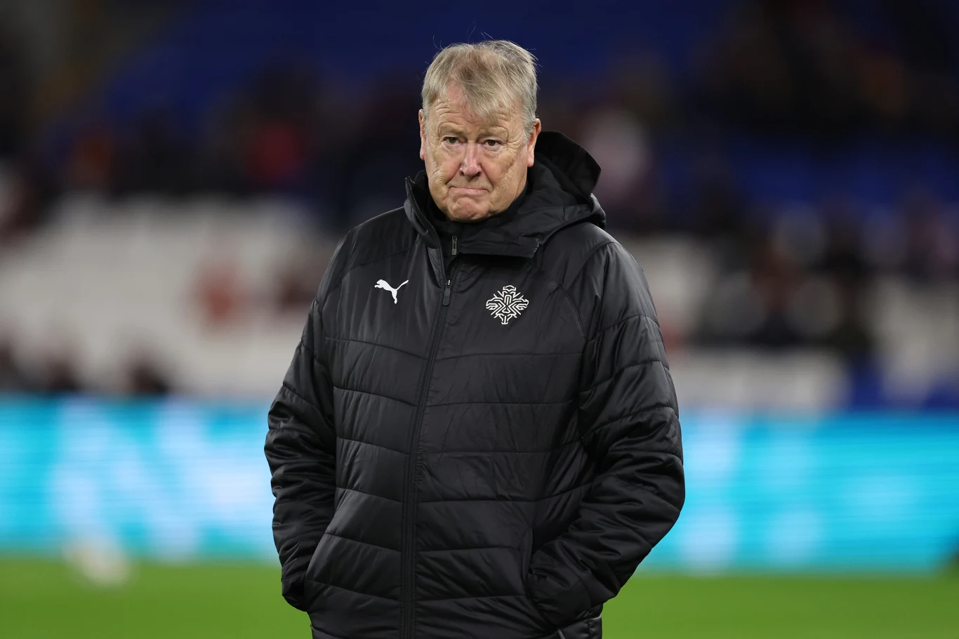 Age Hareide Starszy mężczyzna ubrany w czarną kurtkę sportową z rękoma w kieszeniach, stoi na tle rozmytej trybuny stadionowej.