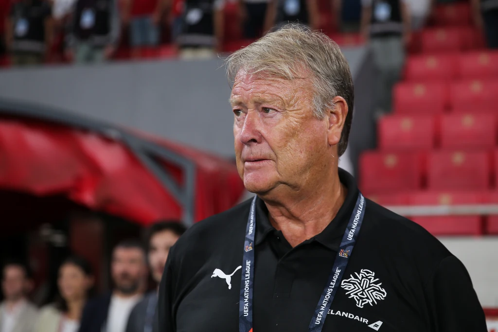 Starszy mężczyzna w sportowej koszulce z logo oraz naszywką z napisem UEFA Islandia, stojący na tle trybun stadionu podczas wydarzenia sportowego, w otoczeniu kilku innych osób niewyraźnie widocznych w tle.