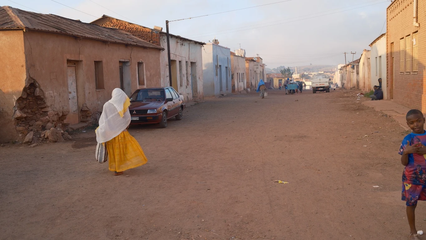 Erytrea Pusta, szeroka ulica z niskimi, zniszczonymi budynkami, po lewej stronie kobieta w białym welonie i żółtej spódnicy, po prawej stronie chłopiec w kolorowej koszulce, kilka osób w oddali, atmosfera codziennego życia w ubogiej dzielnicy.