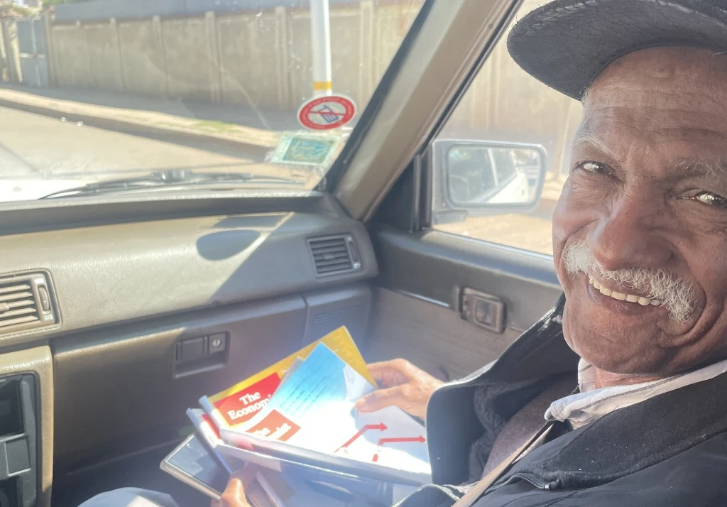 Starszy mężczyzna uśmiecha się do aparatu, siedząc na siedzeniu pasażera w samochodzie i trzymając w ręku kolorowe czasopisma i dokumenty.