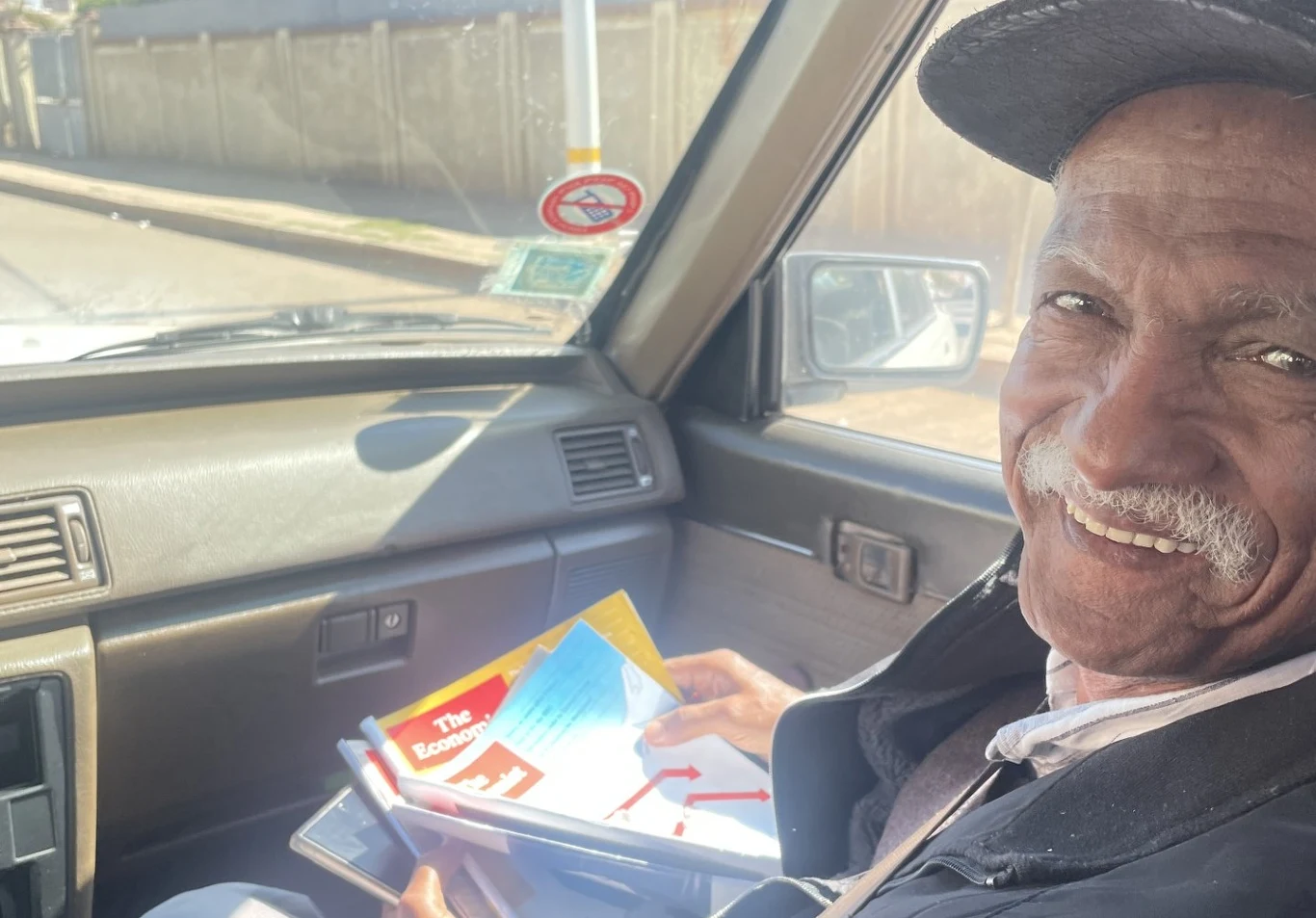 Pan Tekeste. Asmara Starszy mężczyzna uśmiecha się do aparatu, siedząc na siedzeniu pasażera w samochodzie i trzymając w ręku kolorowe czasopisma i dokumenty.