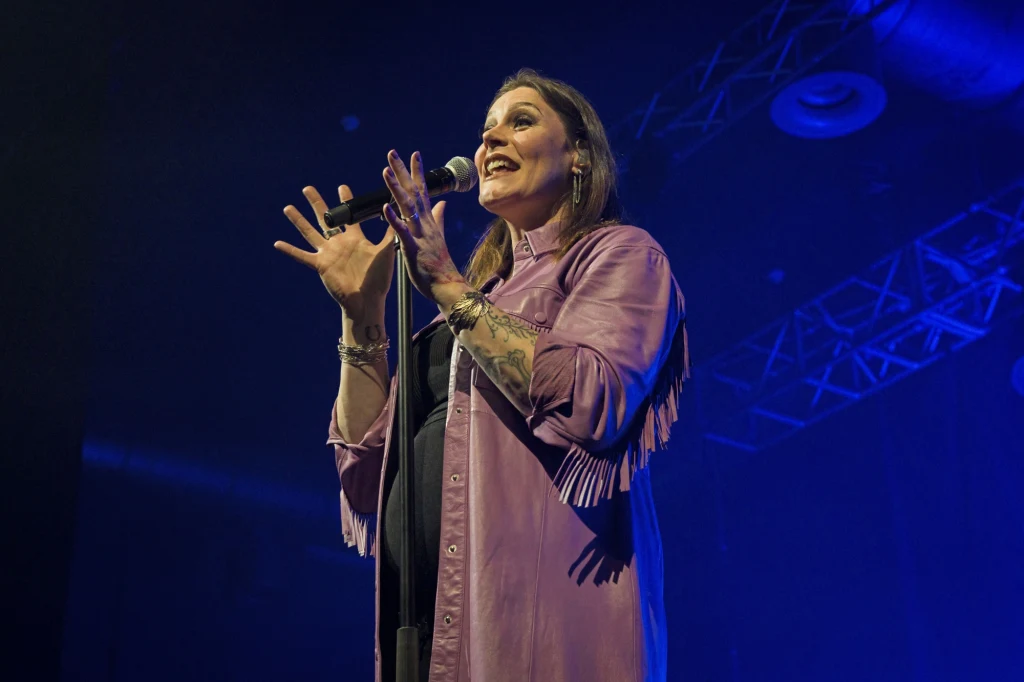 Floor Jansen przyjedzie na Pyrkon do Poznania Kobieta ubrana w fioletową koszulę śpiewa na scenie, trzymając mikrofon w dłoni, w tle widoczne oświetlenie sceniczne i elementy konstrukcji sufitowej.