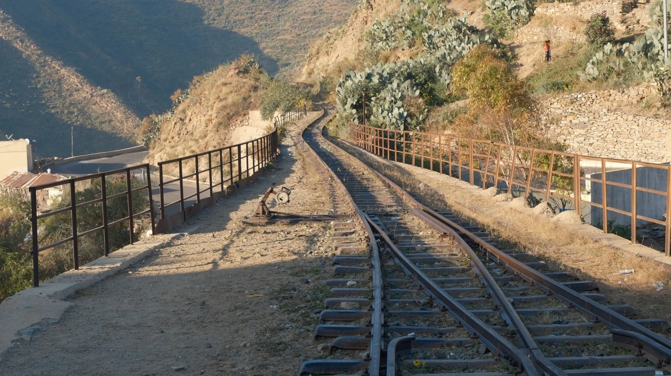 Erytrea Tor kolejowy prowadzący przez górzysty teren z metalowymi barierkami zabezpieczającymi po obu stronach, otoczony roślinnością i pagórkami skąpanymi w świetle słonecznym