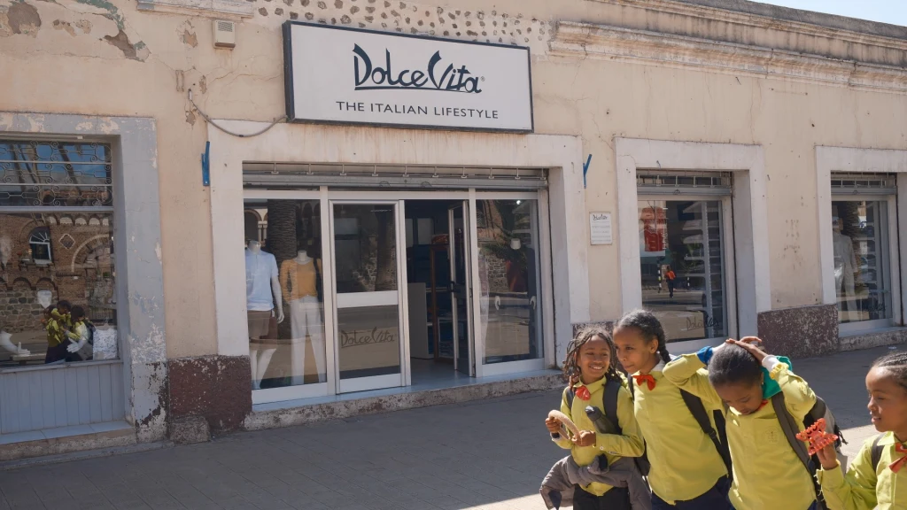 Grupa dzieci w żółtych mundurkach idzie chodnikiem obok zniszczonego budynku z widocznym szyldem sklepu Dolce Vita, za szybą wystawa z ubraniami, jasny dzień i budynek noszący ślady zniszczeń.