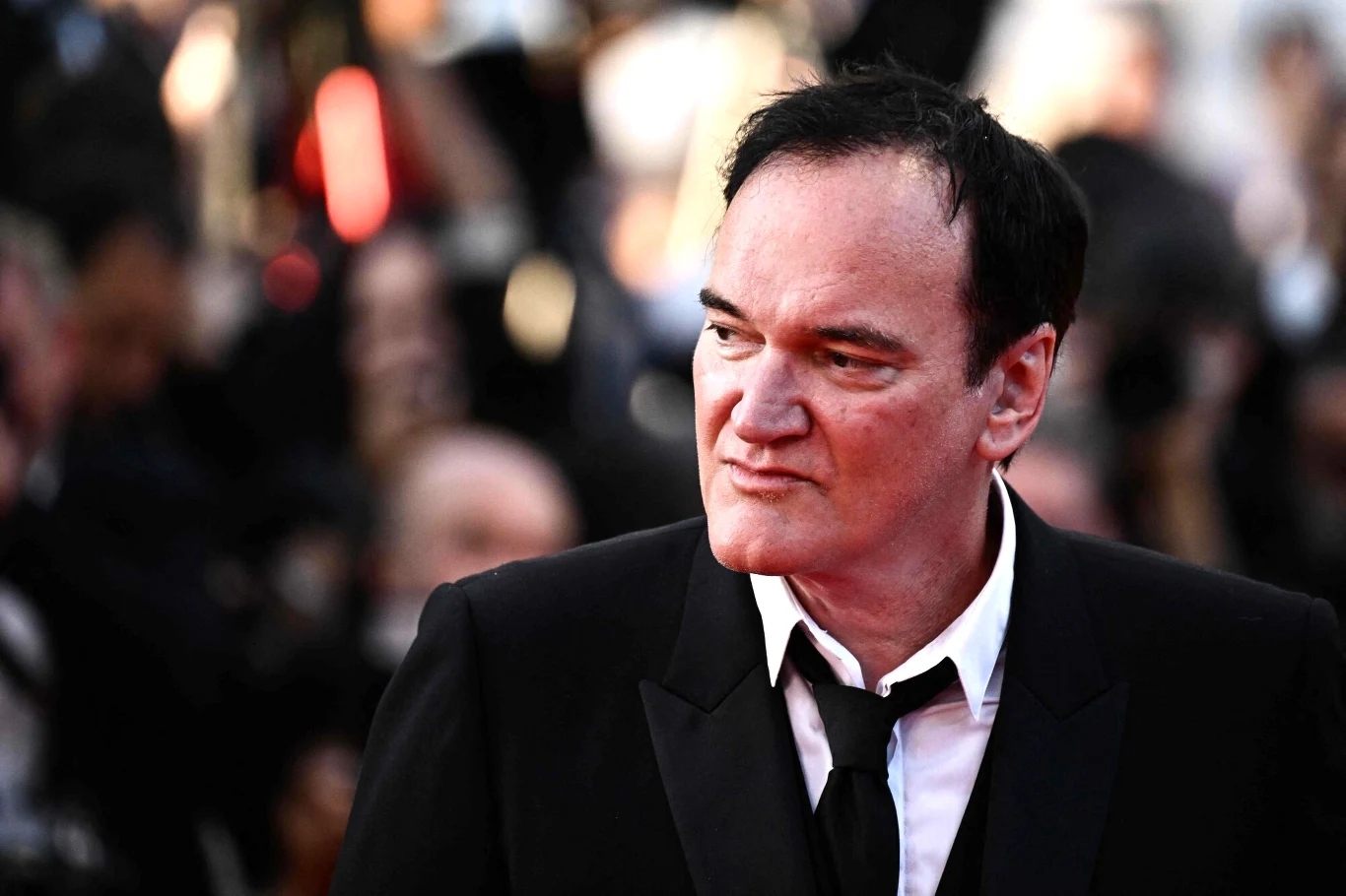Quentin Tarantino Mężczyzna w czarnym garniturze i białej koszuli podczas gali, stoi przed rozmytym tłumem, zwrócony lekko w bok.