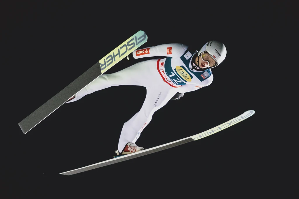 Kacper Tomasiak w trakcie zawodów Pucharu Świata w skokach narciarskich Skoczek narciarski w białym kombinezonie olimpijskim podczas lotu w powietrzu na tle czarnego nieba, z widocznymi nartami i kaskiem ochronnym.