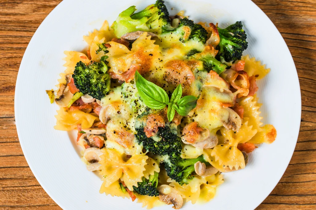 Makaron kokardki z warzywami takimi jak brokuły i pieczarki, posypany stopionym serem oraz udekorowany świeżą bazylią, podany na białym talerzu na drewnianym stole.