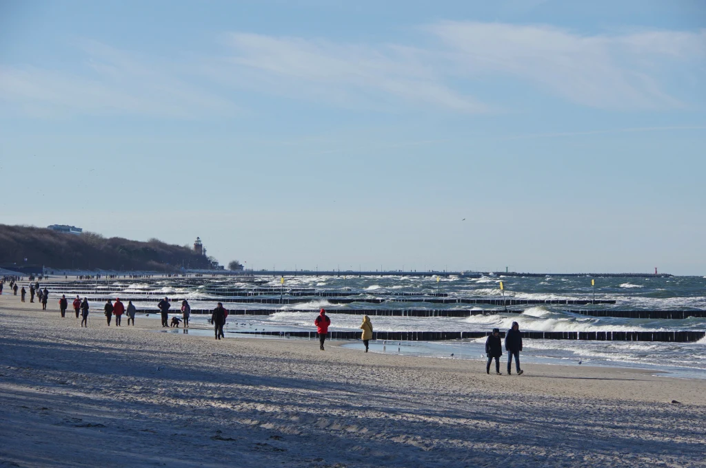 Spacerujące osoby w ciepłych ubraniach na szerokiej, piaszczystej plaży nad morzem z widocznymi falami i odsłoniętymi falochronami, w tle zarys lasu i daleka panorama latarni morskiej.
