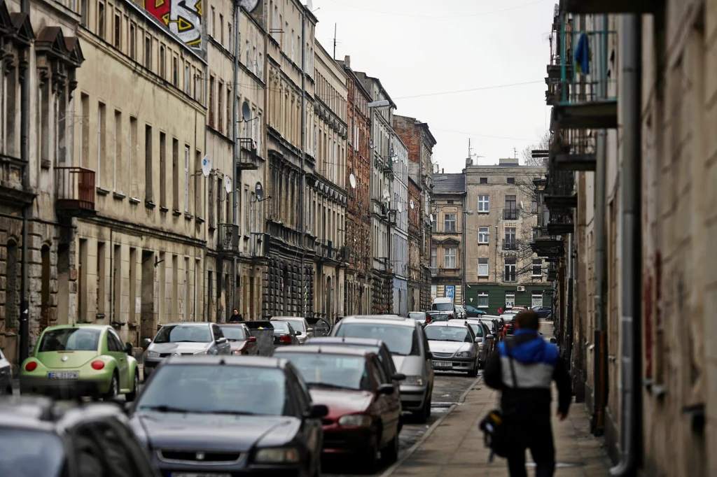 Wąska uliczka miejskiej zabudowy z zabytkowymi, wielopiętrowymi kamienicami po obu stronach, liczne zaparkowane samochody i pojedynczy pieszy idący chodnikiem.