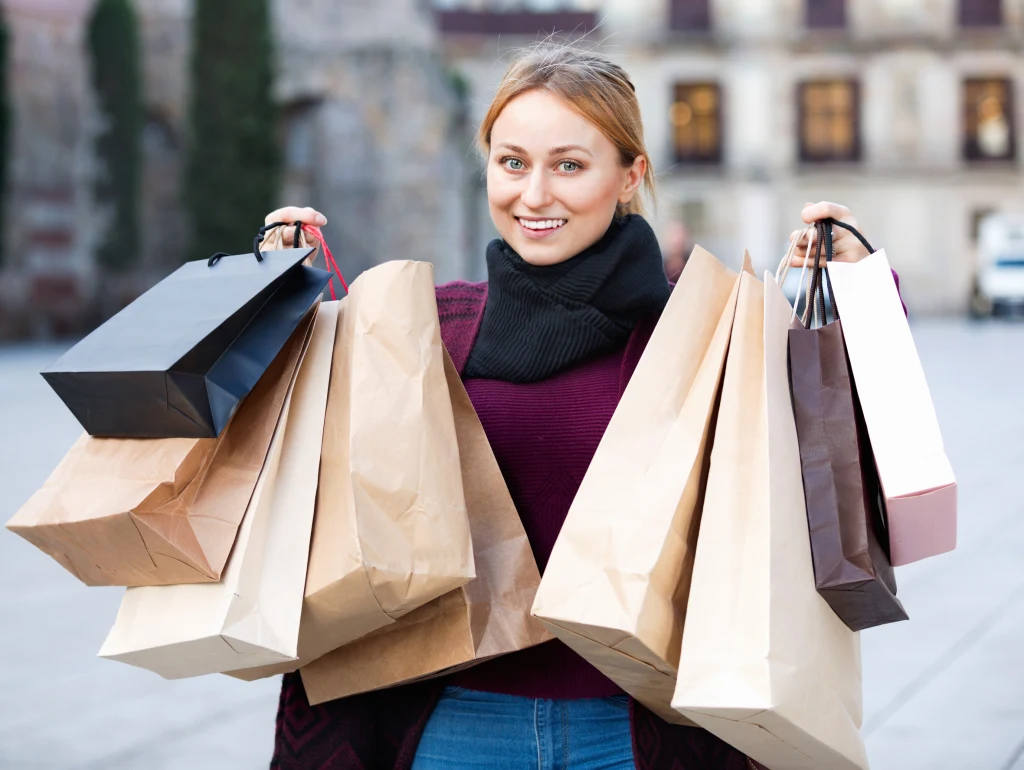 Grudniowy przypływ finansów. Zobacz, kto może liczyć na niespodzianki Uśmiechnięta młoda kobieta trzymająca w obu rękach wiele papierowych toreb zakupowych, ubrana w bordowy sweter i czarny szalik, w tle zabudowa miejska w lekko rozmytym stylu.