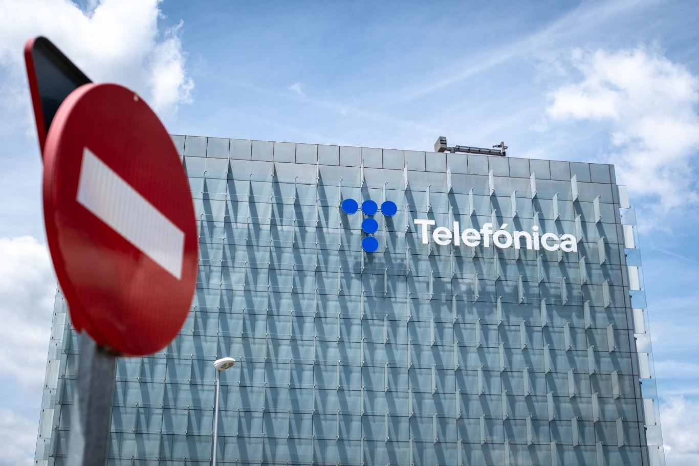 Telefónica zwiększa liczbę zwolnień w planie redukcji zatrudnienia do ponad 5 tys. osób Budynek biurowy o nowoczesnej szklanej fasadzie z widocznym logotypem Telefónica, na pierwszym planie znak zakazu wjazdu na tle błękitnego nieba z chmurami.