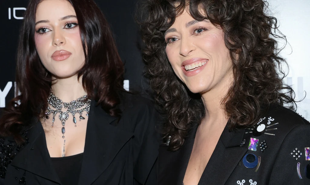 Dwie kobiety w eleganckich, czarnych ubraniach stoją obok siebie na tle z elementami graficznymi, jedna z nich ma kręcone włosy i szeroki uśmiech, druga ma proste, ciemne włosy i na szyi bogato zdobiony naszyjnik.