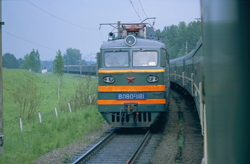 Elektryczna lokomotywa z czerwonym symbolem gwiazdy na przodzie jedzie po torach kolejowych, otoczonych zielonym krajobrazem i drzewami, po lewej stronie widoczny ogrodzony teren.