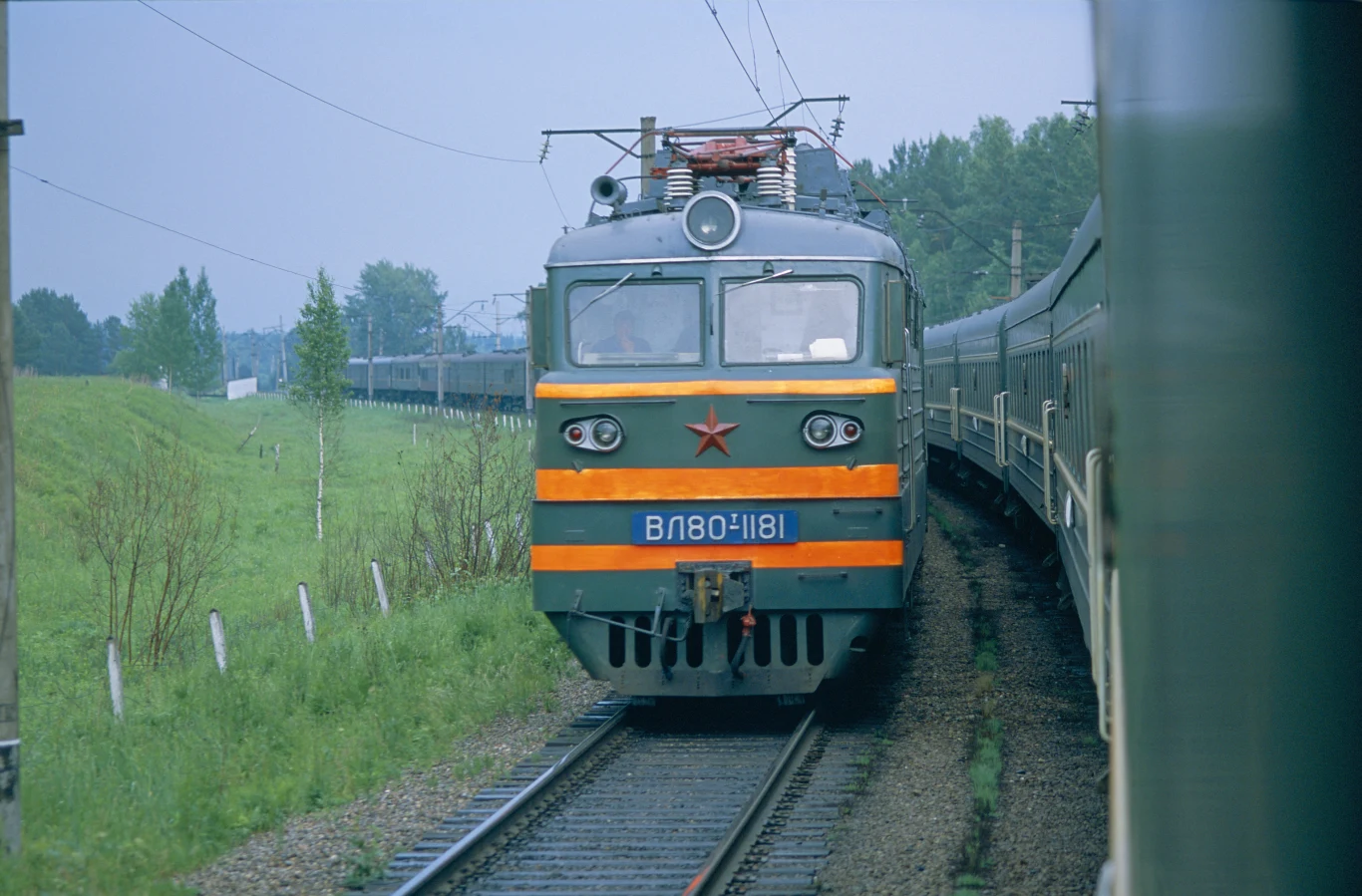 Kreml próbuje ratować znajdujące się w fatalnej sytuacji Koleje Rosyjskie Elektryczna lokomotywa z czerwonym symbolem gwiazdy na przodzie jedzie po torach kolejowych, otoczonych zielonym krajobrazem i drzewami, po lewej stronie widoczny ogrodzony teren.