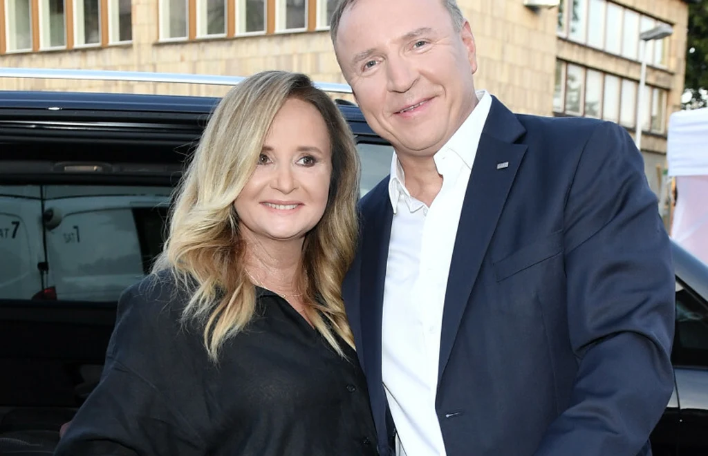 Kobieta o blond włosach w czarnej koszuli oraz mężczyzna w granatowej marynarce i białej koszuli stoją obok siebie uśmiechając się, w tle budynek z dużymi oknami.