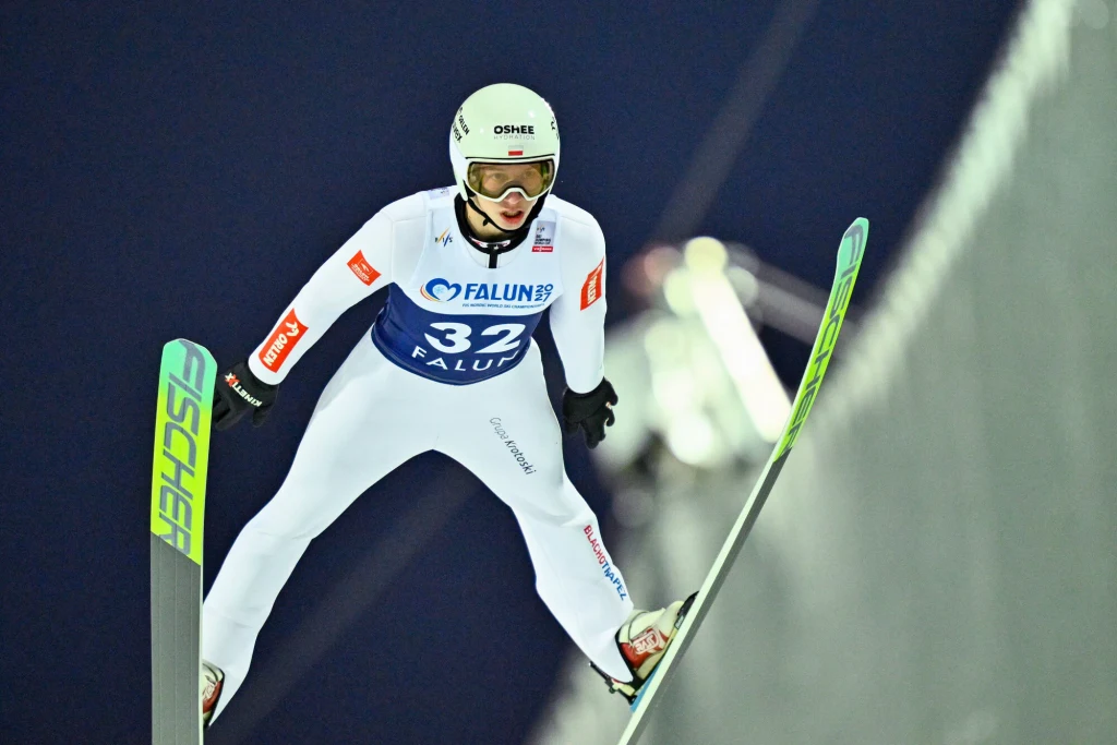 Kacper Tomasiak Skoczek narciarski w białym kombinezonie i kasku w locie podczas konkursu, widoczny numer startowy 32 i napisy na kombinezonie, ciemne tło podkreślające dynamikę skoku.