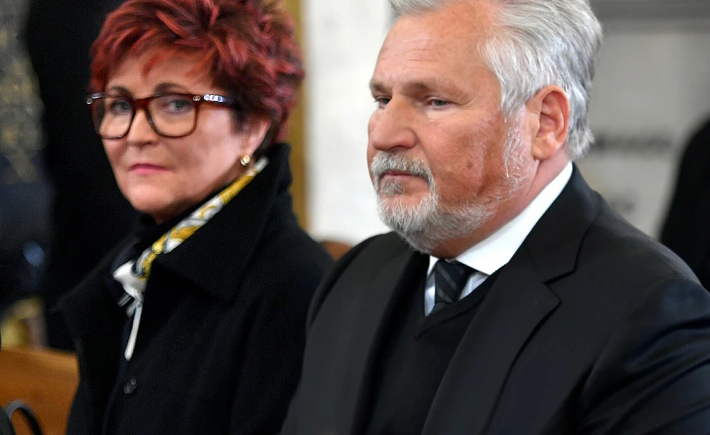 Dwoje elegancko ubranych starszych osób siedzi obok siebie w pomieszczeniu o oficjalnym charakterze, mężczyzna z siwymi włosami i brodą nosi ciemny garnitur i białą koszulę, kobieta w okularach ma krótkie, rude włosy oraz ciemny płaszcz.